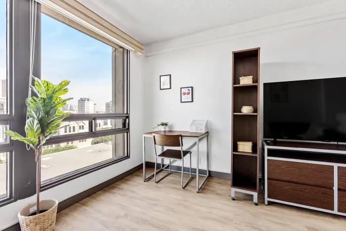 Bright urban apartment corner with large windows, elegant desk, TV, and cozy decor, offering a stylish and functional workspace.